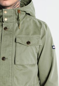 Zelená bunda s kapucí z texturované látky. Má zlaté kovové doplňky, přední kapsy a malou logo na rukávu.