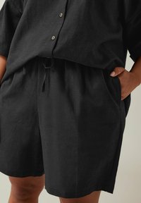 Conjunto corto de lino negro que incluye una camisa de botones holgada y pantalones cortos con cintura elástica, bolsillos laterales y un detalle de cordón.