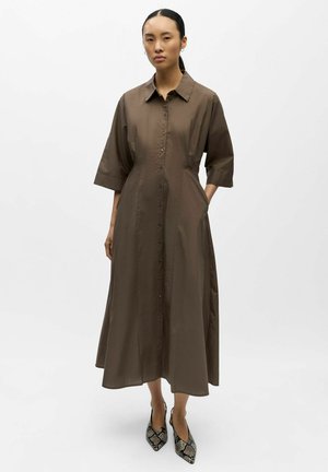 Frau mit langen dunklen Haaren trägt ein braunes Midi-Kleid mit Knopfleiste, Ellenbogen langen Ärmeln und spitz zulaufenden High Heels mit Schlangenledermuster.