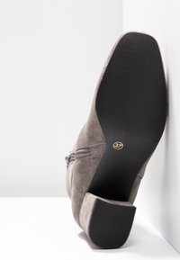 Bottine grise en daim avec une semelle en caoutchouc noire texturée, un petit talon et une fermeture éclair latérale. Taille indiquée : 37.