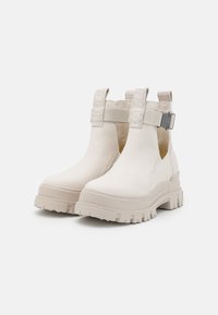 Bottes blanches épaisses jusqu'à la cheville, finition en cuir lisse, panneaux latéraux élastiques, et une boucle métallique décorative sur la bride, avec une semelle robuste.