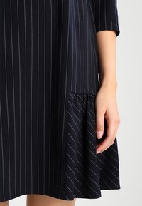Marineblaues Kleid mit vertikalen weißen Streifen, kurzen Ärmeln und einem ausgestellten Saum; strukturierter Stoff mit einem Raffungselement an der unteren Seite.
