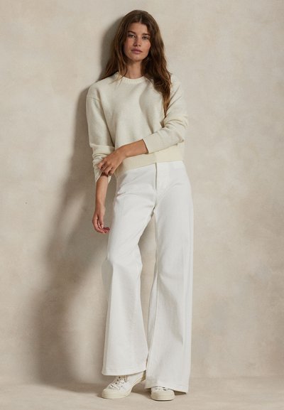 Donna con lunghi capelli castani che indossa un maglione crema, pantaloni bianchi a gamba larga e scarpe da ginnastica bianche, in piedi davanti a uno sfondo beige.