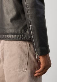 Schwarze Lederjacke mit seitlichem Reißverschluss und glatter Oberfläche, kombiniert mit hellbeigen Stoffhosen mit aufgesetzter Tasche.