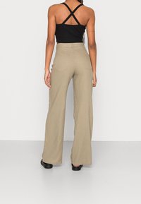 Jasno-beżowe, szerokie spodnie lniane z wysokim stanem i dwoma tylnymi kieszeniami. Modelka nosi czarny, krótki top na ramiączkach z krzyżującymi się paskami.