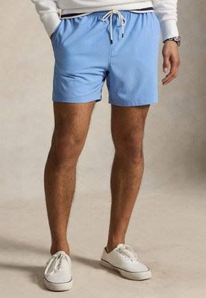 Homme portant un short bleu clair à cordon, des baskets blanches et un haut blanc à manches longues avec un détail aux poignets rayés, debout les mains dans les poches.