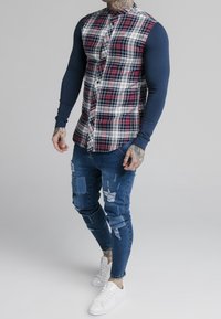 Checked shirt with navy long sleeves, featuring a button-up front and a plaid pattern in red, white, and blue. Worn with distressed blue jeans and white sneakers.