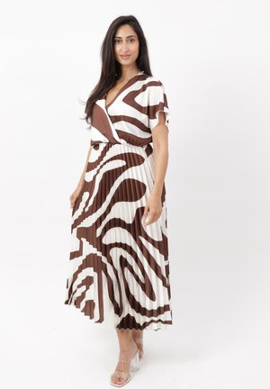 Mujer de pie con un vestido midi plisado con patrón abstracto en blanco y marrón, mangas cortas y tacones de tira transparente.