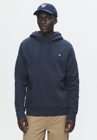 Marineblå hættetrøje med snøre, kengurulomme, ribbede manchetter og et lille logo på brystet. Kombinationen er med beige bukser.