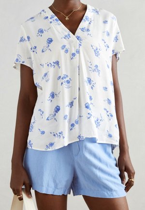 Kvinde iført en hvid kortærmet bluse med blå blomsterprint og lyseblå shorts, accessorized med en guldkæde og ringe.