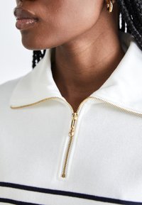 Maglione bianco con zip, caratterizzato da una texture a coste, con una zip dorata e due strisce orizzontali navy vicino all'orlo. Design con colletto ampio.