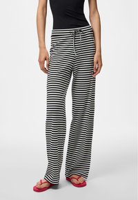 Henkilö, jolla on päällään mustavalkoiset vaakaraitaiset leveälahkeiset housut, joissa on nyörivyötärö, ja punaiset varvastossut yksiväristä taustaa vasten.