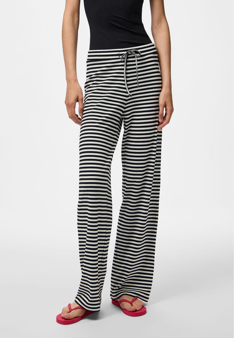 Henkilö, jolla on päällään mustavalkoiset vaakaraitaiset leveälahkeiset housut, joissa on nyörivyötärö, ja punaiset varvastossut yksiväristä taustaa vasten.