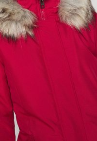 Czerwona kurtka parka z miękkim sztucznym futerkiem na kołnierzu, wyposażona w zamek błyskawiczny i widoczne przeszycia na materiale.