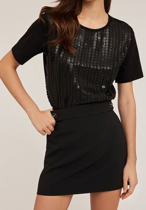 Woman wearing a black short-sleeve top with textured front and a high-waisted black skirt with side button details.