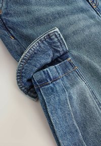 Jeans din denim cu un finisaj albastru deschis, având o manșetă răsucită și cusături în contrast. Țesătura are un aspect texturat, cu modele de țesătură vizibile.