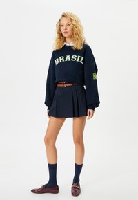 Marineblaues Sweatshirt mit der Aufschrift "BRASIL" in grünen Buchstaben, kombiniert mit einem dunklen, gefalteten Rock, marineblauen Kniestrümpfen und burgunderfarbenen Slippern.
