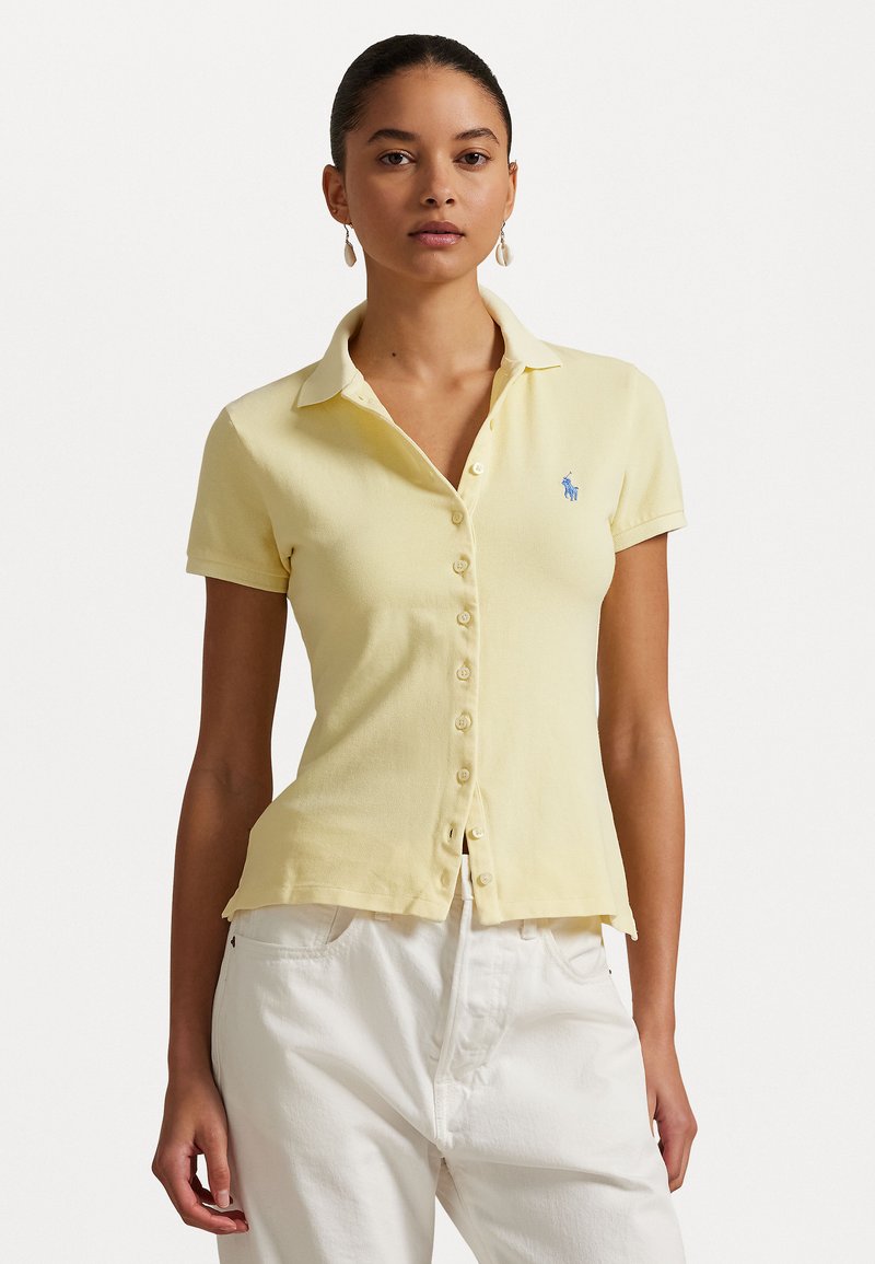 Dzeltena polo krekla izgatavota no teksturēta auduma, ar pogu aizdari priekšā, īsām piedurknēm un mazu zilu logo kreisajā krūtī.