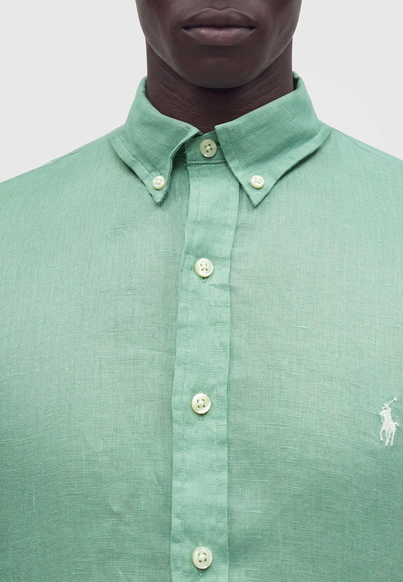 Gros plan d'une personne portant une chemise en lin vert clair boutonnée avec un col pointu et un petit logo blanc brodé sur la poitrine.