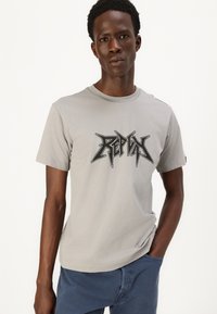 Grå t-shirt med korte ærmer, der har et sort grafisk logo i en skarp stil. Glat tekstur, rund hals design, parret med blå bukser.