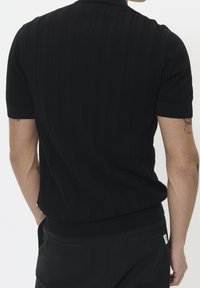 Schwarzes Polohemd mit kurzen Ärmeln, das ein strukturiertes Muster, einen gerippten Kragen und Bündchen sowie eine lockere Passform aufweist. Rückseite des Hemds gezeigt.
