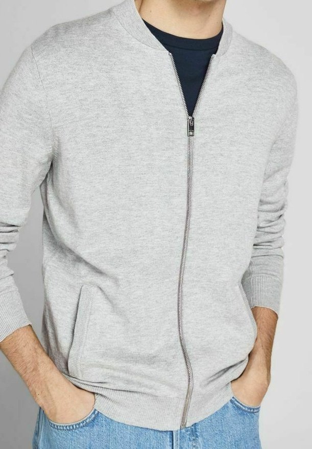 Man som bär en ljusgrå zip-up kofta över en mörk skjorta, med händerna i fickorna på ljusblå jeans.