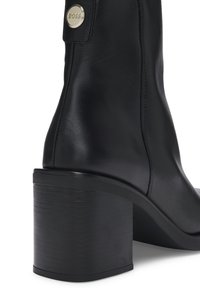 Botín de cuero negro hasta el tobillo con tacón de bloque, textura suave y punta redonda. Decorado con un acento metálico del logo cerca de la parte superior.