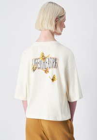 La camiseta oversized de color crema presenta mariposas impresas y el texto "La vida es un tesoro" en la parte trasera. Mangas cortas, tejido suave, ajuste relajado.