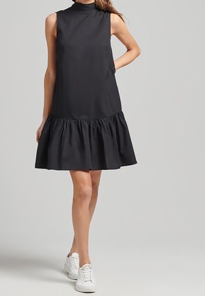 Frau trägt ärmelloses schwarzes Kleid mit tiefer Taille und Rüschensaum sowie weißen Turnschuhen, steht vor einfarbigem grauem Hintergrund.