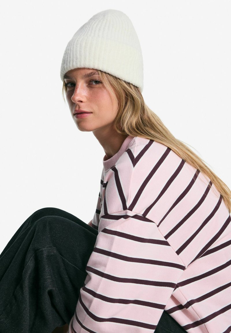 Beanie alb cu model ribatat purtat cu un pulover cu dungi roz și închise. Subiectul este așezat, cu părul lung și pantaloni negri, pe un fundal alb.