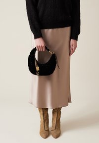 Black, textured handbag with a rounded shape, gold hardware accents, and a short handle, held by a person wearing a black sweater and satin skirt.