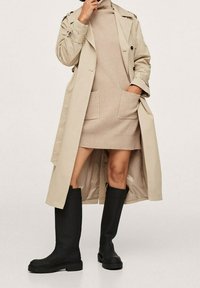 Trench coat beige su un vestito beige a coste con due tasche frontali, abbinato a stivali neri in gomma alti con suole testurizzate.