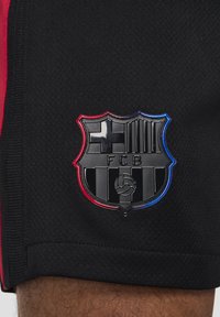 Sorte atletiske shorts har et tekstureret stof med en kontrastfarvet rød sidestribe og et forhøjet sort skjoldformet logo med farveaccentuer.