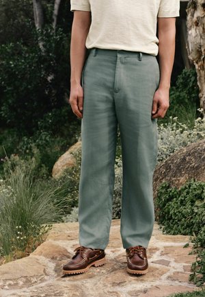 Persona che indossa una camicia a maniche corte beige chiaro, pantaloni verdi larghi e scarpe di pelle marrone, in piedi su un sentiero di pietra in un giardino.