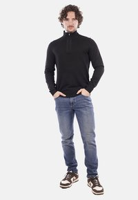 Pullover nero con colletto a zip, indossato con jeans blu e sneakers marroni. Caratterizzato da un design aderente e una texture liscia. Stile casual.