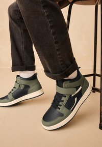 Baskets montantes en cuir vert olive et bleu marine, avec une finition texturée, une sangle ajustable et une semelle blanche.