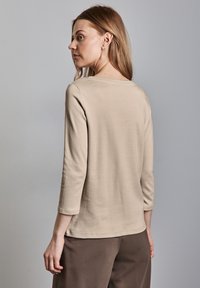 Beige Langarm-T-Shirt mit lockerer Passform, Rundhalsausschnitt und weichem Baumwollstoff, kombiniert mit braunen Hosen, von hinten betrachtet.