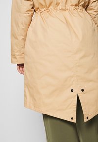 Light beige, mid-length coat with elastic waistband, side slits, and black snap buttons at the hem. Smooth texture, minimal design details.