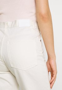 Designer de ganga branca com uma cintura alta, apresentando dois bolsos traseiros e uma placa de couro simples na parte de trás da cintura. Textura suave.