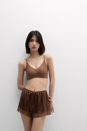 Frau mit schulterlangem schwarzen Haar, die einen braunen Bralette und passende Satin-Shorts mit Spitzenbesatz trägt, stehend vor einem einfarbigen weißen Hintergrund.