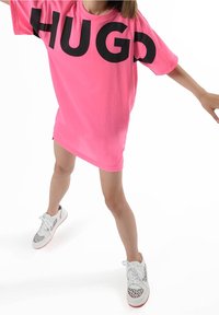 Maglietta oversize rosa con la scritta "HUGO" in lettere nere e spesse sul davanti. Indossata con sneaker bianche caratterizzate da un motivo grafico nero.