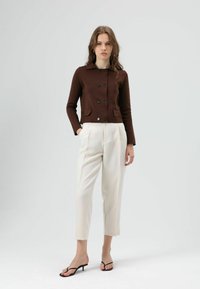 Bruin kort vest met knoopsluiting, twee zakken aan de voorkant, gecombineerd met crèmekleurige taps toelopende broek en zwarte sandalen met hak. Gladde textuur.