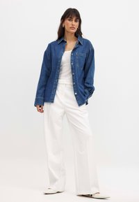 Camisa de mezclilla en azul, con cuello puntiagudo, cierre de botones y un bolsillo en el pecho, llevada sobre una blusa blanca y pantalones de pierna ancha blancos.