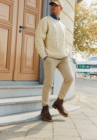Stivali da trekking color tan, pantaloni beige e un maglione con lavorazione a trecce color crema. L'ambientazione presenta una porta marrone chiaro e gradini in pietra grigia.