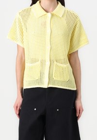 Model v krátkém rukávu, žluté pletené cardigan s knoflíky a límcem, se dvěma předními kapsami, kombinovaný s černými kalhotami.