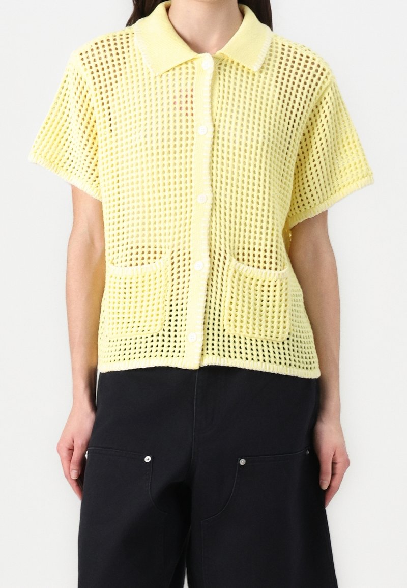 Model v krátkém rukávu, žluté pletené cardigan s knoflíky a límcem, se dvěma předními kapsami, kombinovaný s černými kalhotami.