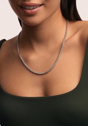 Collar de cadena de plata con diseño trenzado, llevado sobre una blusa verde oscuro. Textura suave con acabado reflectante.