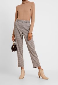 Pull col roulé beige avec poignets côtelés, pantalon taille haute à carreaux, et bottines beige clair à fermeture éclair latérale. Accessoire sac à main marron.