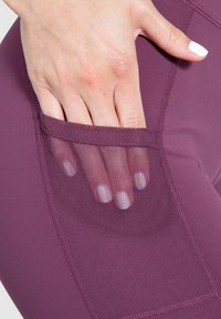 Burgundy leggings dengan saku samping jaring. Kainnya elastis dan halus, menunjukkan tangan yang meraih ke dalam saku.