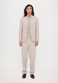 Costume en lin beige clair comprenant un blazer ajusté avec des revers à cran, un pantalon assorti et une chemise grise à boutons en dessous. Des chaussures foncées complètent le look.